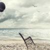 Surreal image of a chair held in balance by flying balloons on a beach